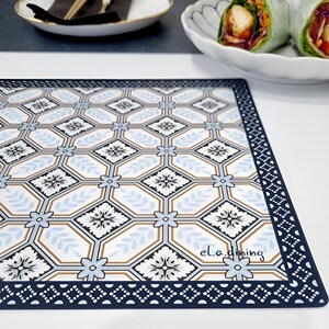 May include: A rectangular placemat with a geometric pattern in white, light blue, and brown, framed by a navy blue border. The design features floral and leaf motifs within hexagonal shapes. The text "el.a.dining" is visible in the lower right corner.