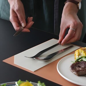 Peut inclure: Un ensemble de fourchette, cuillère et couteau en acier inoxydable sur un set de table blanc et pêche. Les couverts sont placés à côté d'une assiette de nourriture, dont un steak et du maïs en épi. La scène se déroule sur une table noire.