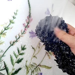 May include: A close-up of a floral-patterned surface being cleaned with a dark blue, textured sponge. The surface features watercolor-style illustrations of various flowers and greenery in shades of purple, green, and yellow. Water droplets are visible on the surface.