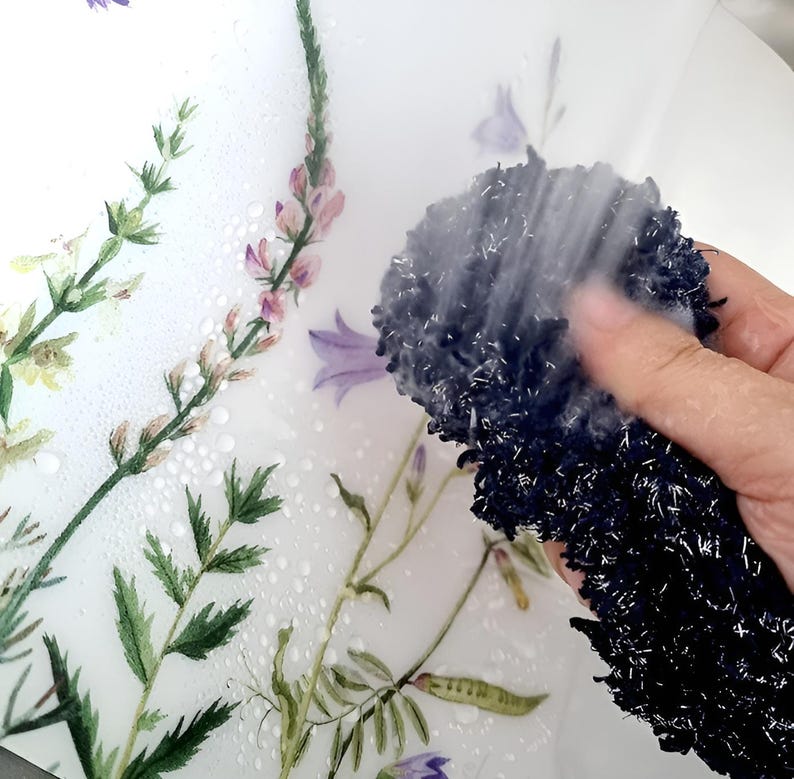 May include: A close-up of a floral-patterned surface being cleaned with a dark blue, textured sponge. The surface features watercolor-style illustrations of various flowers and greenery in shades of purple, green, and yellow. Water droplets are visible on the surface.