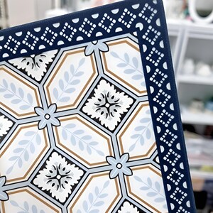 May include: Close-up of a decorative square tile with a geometric pattern. The tile features a white background with light blue, brown, and black floral and leaf designs. A dark blue border with a repeating white pattern frames the tile.