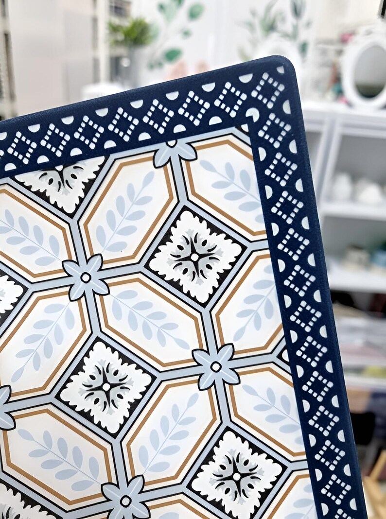 May include: Close-up of a decorative square tile with a geometric pattern. The tile features a white background with light blue, brown, and black floral and leaf designs. A dark blue border with a repeating white pattern frames the tile.