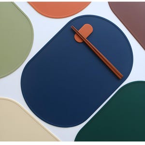 May include: A selection of colourful placemats in shades of blue, green, and orange. Wooden chopsticks rest on an orange chopstick rest on a blue placemat. The placemats are oval-shaped, and the image is taken from above.