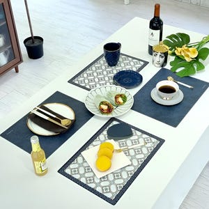 May include: A table setting with various items, including a bottle of wine, a cup of coffee, plates with food, and decorative placemats. The placemats are dark blue with a geometric pattern. The table is white.