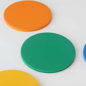 May include: A collection of colorful, circular coasters in orange, green, blue, and yellow. The coasters are arranged on a white surface, with each coaster's solid color clearly visible. The design is simple and modern.