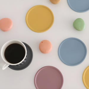 May include: Overhead shot of a white coffee cup filled with black coffee, resting on a dark gray saucer. Surrounding the cup are several colorful, round coasters in shades of yellow, blue, pink, green, and purple. Also visible are small, round, peach-colored objects.
