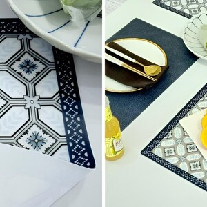 May include: A set of decorative placemats with a geometric pattern in white, gray, and navy blue. The placemats feature a border design and are shown with plates, cutlery, and food. The placemats are rectangular.