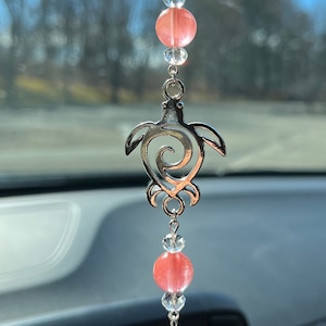 May include: A silver car charm with a pink and clear crystal beaded chain and a silver turtle pendant. The turtle has a swirl design in its shell.
