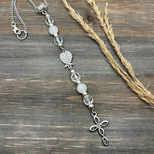 May include: A silver car charm with a white crystal bead and angel wing charm, hanging from a silver chain with a cross at the bottom.