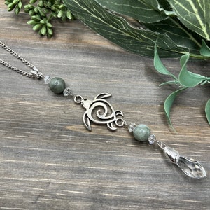 May include: A silver turtle charm with a swirl design hangs from a silver chain with green and clear crystal beads. The charm is hanging against a wood background.