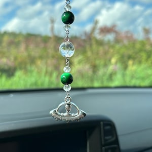 May include: A silver car charm with a UFO design and the text "I want to believe". The charm is hanging from a chain with green and clear crystal beads.