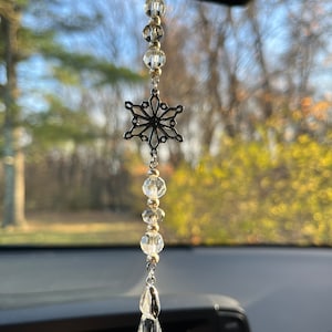 May include: A silver car charm featuring a snowflake pendant, clear crystal beads, and a teardrop-shaped crystal. The charm hangs from a silver chain and is suspended in front of a blurred outdoor background.