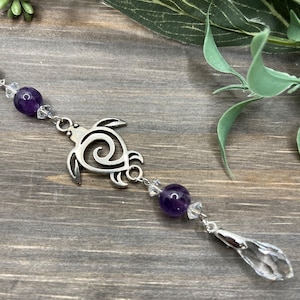 May include: A silver turtle charm with a swirl design hangs from a crystal and amethyst beaded chain. The chain ends with a clear crystal teardrop.