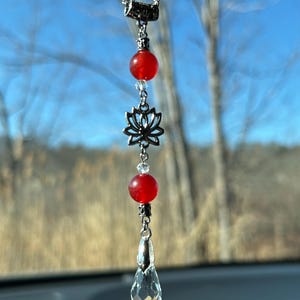 May include: A silver car charm with a chain, featuring a red bead, a silver lotus flower, a clear crystal teardrop, and a small clear bead. The charm hangs against a blurred outdoor background.