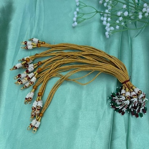 May include: A collection of gold-colored tassels with beaded accents in white, red, and green. The tassels are tied together and arranged on a light green fabric, with white floral accents in the background. The tassels are likely used for decorative purposes.