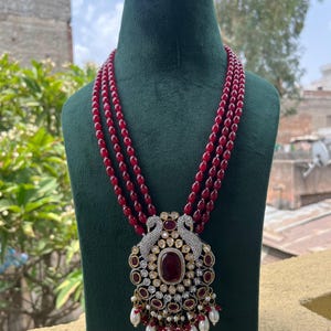 May include: A three-strand red beaded necklace with a large pendant. The pendant features a central oval red stone, surrounded by smaller red stones, clear crystals, and white pearls. A matching bracelet sits above the necklace.