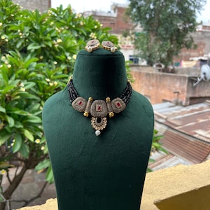 Könnte beinhalten: Ein aufwendiges Halsketten- und Ohrring-Set, präsentiert auf einer grünen Samtbüste. Die Halskette hat schwarze Perlenstränge, goldfarbene Filigranquadrate mit roten Akzenten und einen zentralen Anhänger mit einer Perle. Die Ohrringe spiegeln das Design wider.