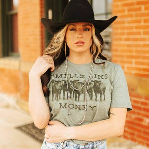 May include: A woman wearing a green t-shirt with a graphic of cows and the text "Smells Like Money". She is also wearing a black cowboy hat, a silver necklace, and blue jeans.