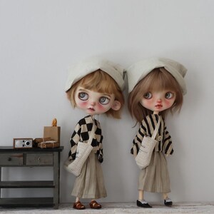 May include: Two dolls dressed in matching outfits stand side-by-side. Each doll wears a beige skirt, a patterned jacket, and a white hat. One jacket has a black and white checkered pattern, and the other has black and white stripes. They each carry a white bag.