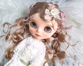 May include: A doll with long, curly, light brown hair styled in braids with bows. The doll is wearing a white dress with a floral embroidery design and has a delicate, painted face with large, blue eyes.