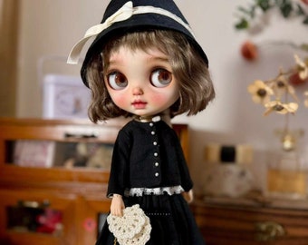 May include: A doll wearing a black dress and jacket with a white lace trim. The doll has brown hair and is wearing a black hat with a white ribbon. The doll is holding a small white crocheted purse with a gold chain.