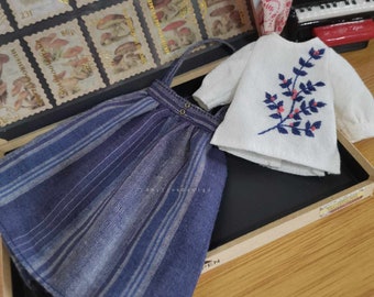 May include: A blue and white striped skirt with a white top featuring an embroidered floral design. The top has long sleeves and a round neckline. The skirt has a simple design with a few buttons on the front.