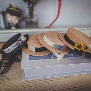 May include: Three straw hats with black ribbon trim and floral embellishments. The hats are arranged on a white box with the text "all or nothing, now or never" and "Falling in love" printed on it.