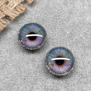 Op de afbeelding: Twee ronde, paarse en blauwe glazen ogen met zilveren glitter rond de randen. De ogen hebben een witte lijn in het midden en een bruine iris.