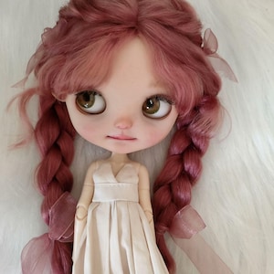 Peut inclure: Une poup&eacute;e &agrave; la peau claire, aux grands yeux bruns et aux joues roses. La poup&eacute;e a de longs cheveux roses tress&eacute;s orn&eacute;s de rubans. Elle porte une robe cr&egrave;me avec un col en V et une taille ajust&eacute;e. L'expression de la poup&eacute;e est douce et sereine.