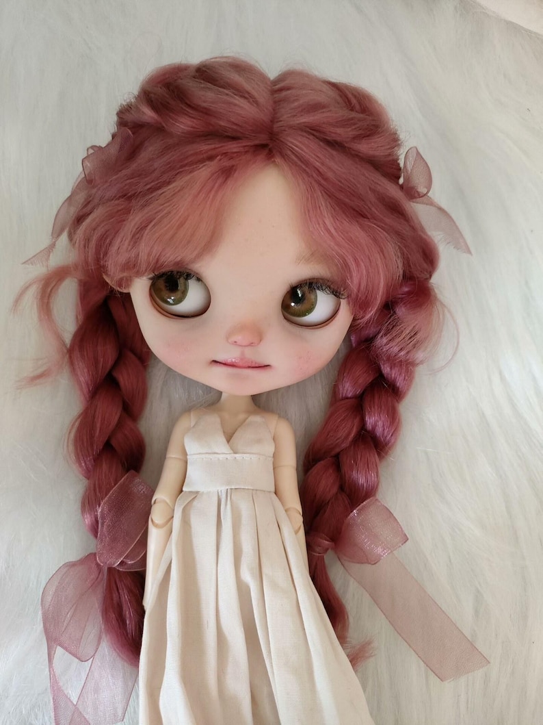Peut inclure: Une poup&eacute;e &agrave; la peau claire, aux grands yeux bruns et aux joues roses. La poup&eacute;e a de longs cheveux roses tress&eacute;s orn&eacute;s de rubans. Elle porte une robe cr&egrave;me avec un col en V et une taille ajust&eacute;e. L'expression de la poup&eacute;e est douce et sereine.