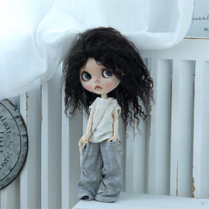 May include: A doll with long, curly black hair wearing a white tank top and gray pants. The doll is sitting on a white chair.