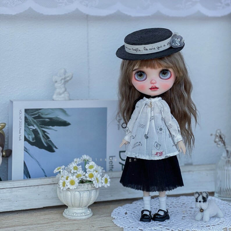 May include: A doll with long brown hair wearing a black and white dress, a black hat with a white ribbon that says "bafle, la vie qui a &eacute;t&eacute;", and black shoes. The doll is standing on a white doily with a white flower pot with white flowers in it.