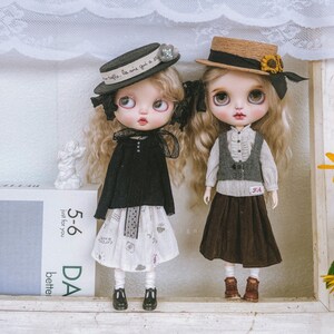 May include: Two dolls with long brown hair wearing different outfits and hats. The doll on the left is wearing a black sweater, a white skirt with a black lace trim, and a black and white hat with the text "la vie qui a du style". The doll on the right is wearing a white and gray striped shirt, a brown vest, a brown skirt, and a brown straw hat with a black ribbon and a brown bow.