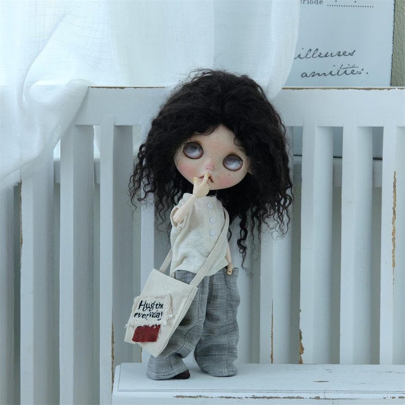 May include: A doll with long black curly hair wearing a white shirt, gray pants, and a canvas bag with the text "Hustle everyday!" embroidered on it.