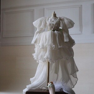 May include: A white, layered, ruffled dress with a long train on a mannequin. The dress has a green sash and is displayed on a wooden box with the text "High & Fine Doll" and "Since" on it. Two doll legs with white socks and green bows are on the box.