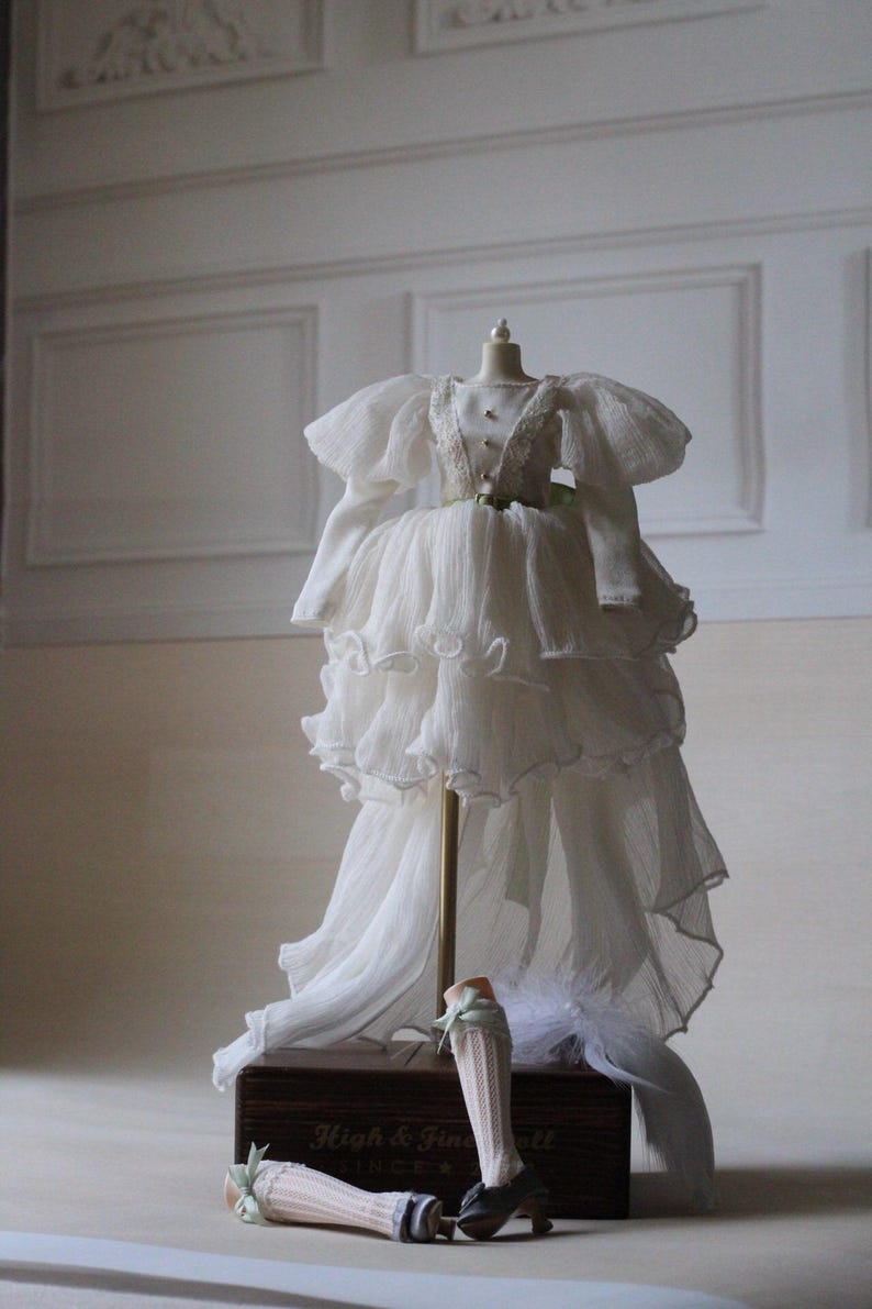 May include: A white, layered, ruffled dress with a long train on a mannequin. The dress has a green sash and is displayed on a wooden box with the text "High & Fine Doll" and "Since" on it. Two doll legs with white socks and green bows are on the box.
