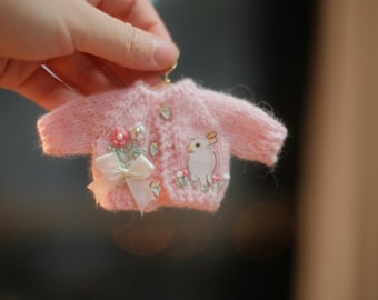 May include: A pink knitted cardigan with a white bunny and floral embroidery. The cardigan has a white bow and pearl-like buttons.