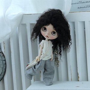 May include: A doll with long, curly black hair wearing a light beige shirt and gray pants. The doll is holding a brown bag and has a necklace with charms.