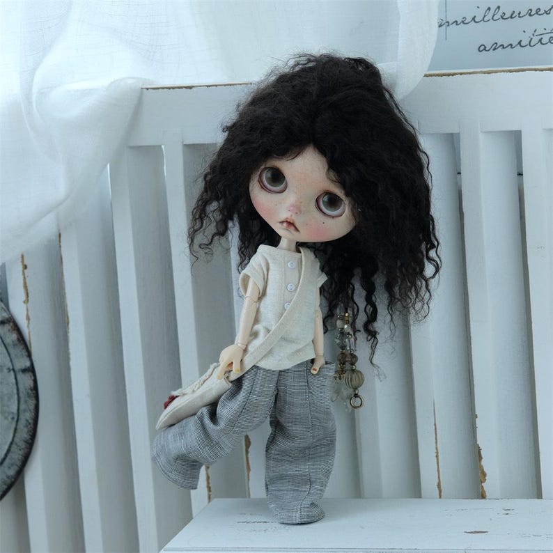 May include: A doll with long, curly black hair wearing a light beige shirt and gray pants. The doll is holding a brown bag and has a necklace with charms.