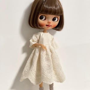 May include: A doll with a bob haircut and a white dress with a floral lace design. The doll is wearing gray socks and black Mary Jane shoes. The doll has large eyes and a small tongue sticking out.