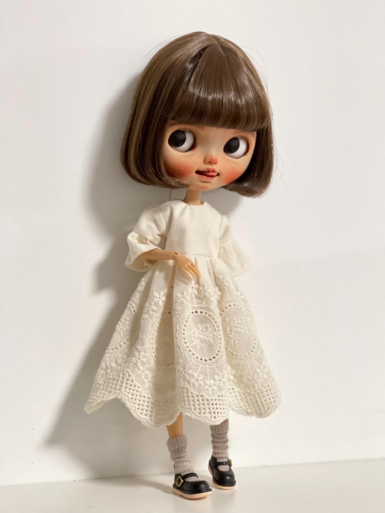 May include: A doll with a bob haircut and a white dress with a floral lace design. The doll is wearing gray socks and black Mary Jane shoes. The doll has large eyes and a small tongue sticking out.