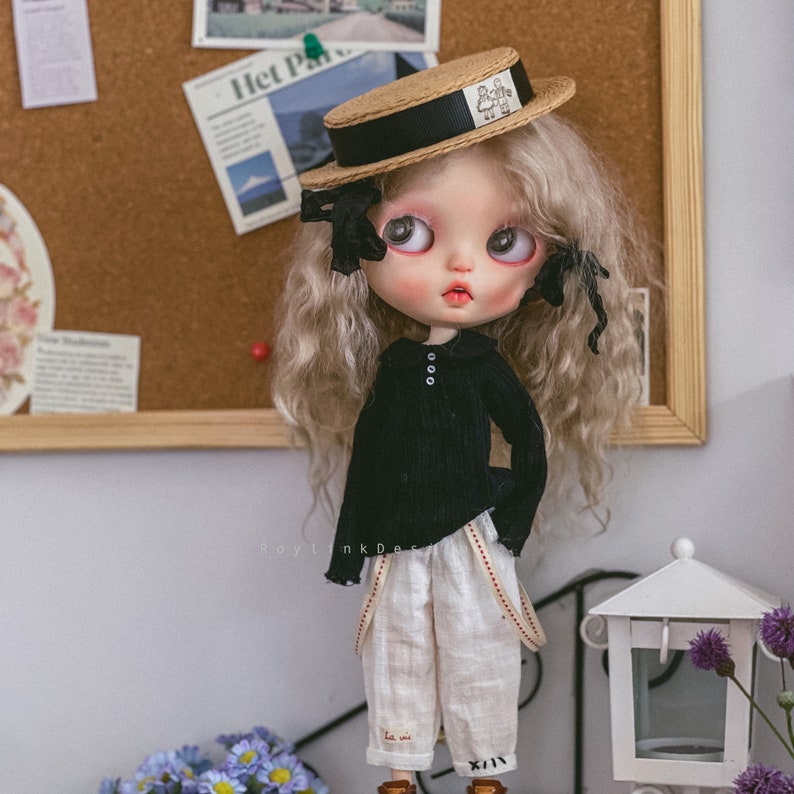 May include: A doll with blonde hair wearing a black sweater, white pants with suspenders, and a straw hat with a black ribbon. The doll has blue eyes and a light pink complexion.