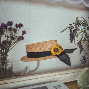 May include: A straw hat with a black ribbon and two yellow sunflower embellishments. The hat is on a white stand.