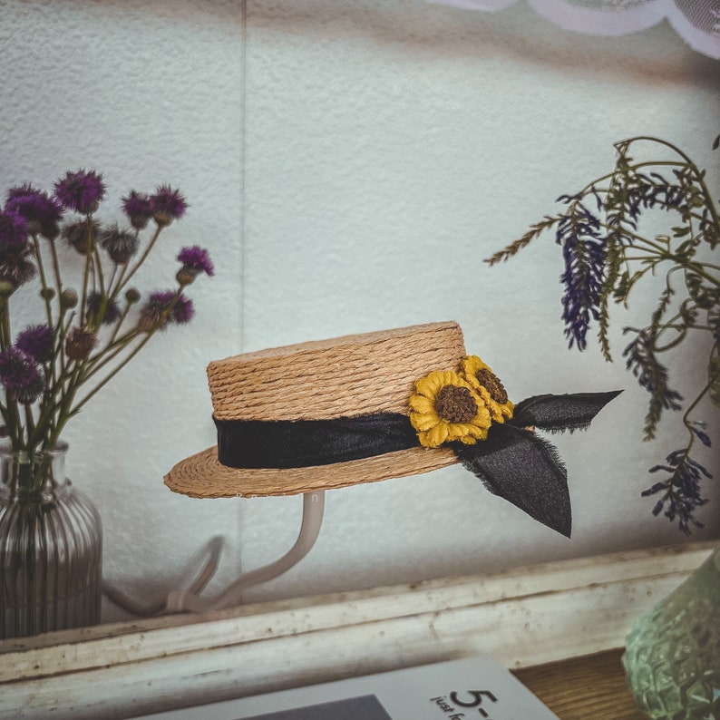 May include: A straw hat with a black ribbon and two yellow sunflower embellishments. The hat is on a white stand.