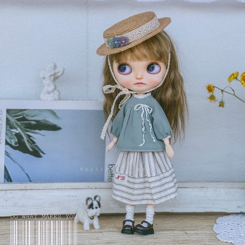May include: A doll wearing a blue and white striped skirt, a light blue blouse with a white bow, and a brown straw hat with a white lace trim. The doll has long brown hair and blue eyes. The doll is standing on a wooden surface with a small white dog figurine in front of it.