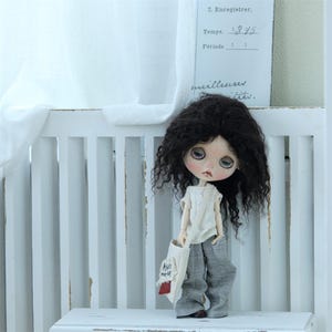 May include: A doll with long, curly black hair, wearing a white shirt and gray pants. The doll is holding a small white bag with red accents and the words "Hello there" written on it. The doll is standing in front of a white wooden fence.