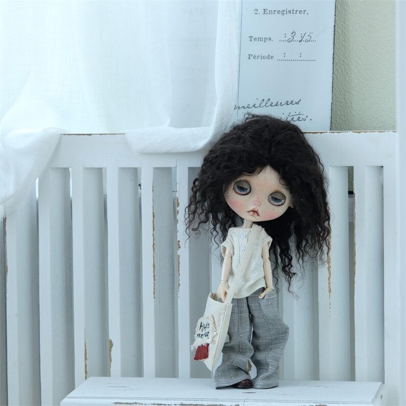 May include: A doll with long, curly black hair, wearing a white shirt and gray pants. The doll is holding a small white bag with red accents and the words "Hello there" written on it. The doll is standing in front of a white wooden fence.