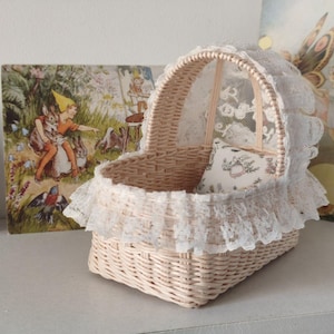 Puede incluir: Una cuna de muñeca de mimbre beige claro con capota y ribete de encaje blanco. La cuna tiene una pequeña almohada estampada en el interior. Se puede ver una obra de arte decorativa en el fondo.