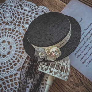May include: A black straw hat with a white ribbon that says "Par un" in French. The hat is decorated with two white fabric flowers with pink and blue accents. The hat is sitting on a white birdcage.