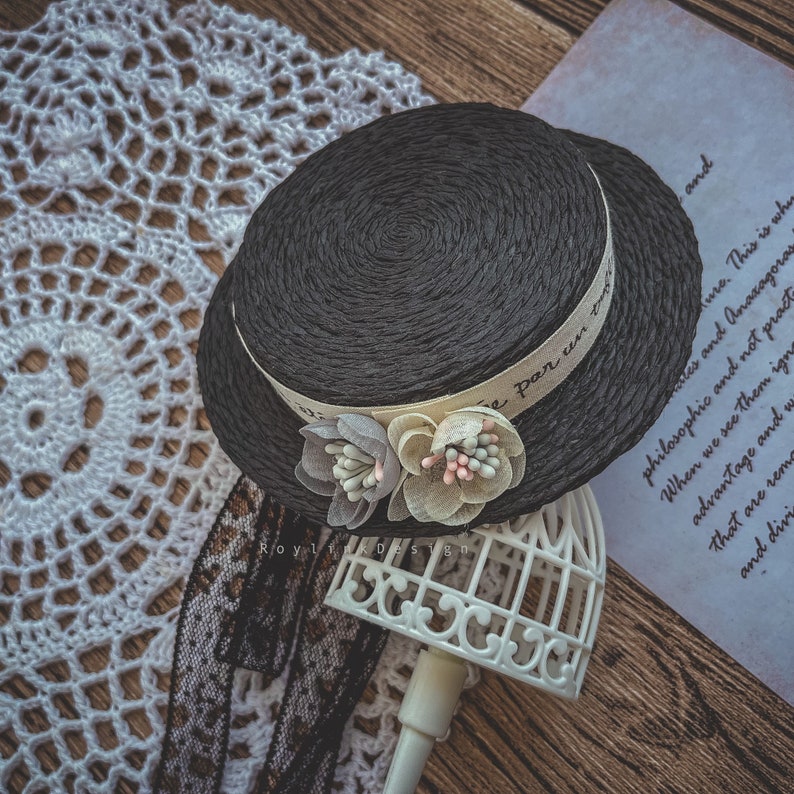 May include: A black straw hat with a white ribbon that says "Par un" in French. The hat is decorated with two white fabric flowers with pink and blue accents. The hat is sitting on a white birdcage.